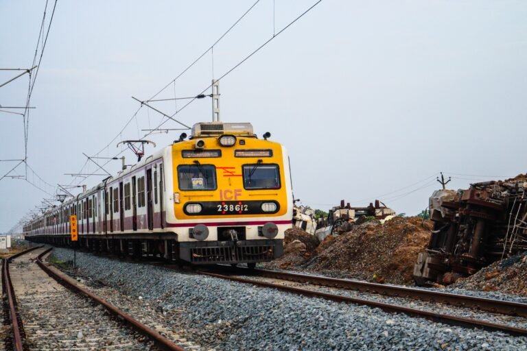 Local commuter train on tracks passing by wreckage, with clear sky backdrop.