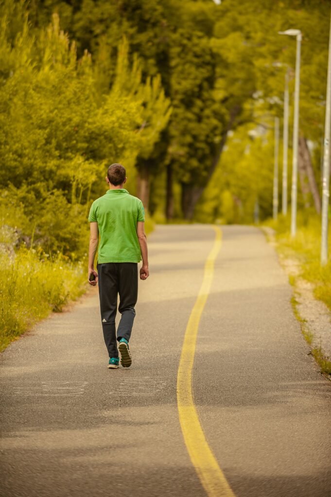 Back view of faceless male in sportswear strolling on asphalt roadway near bright trees in countryside