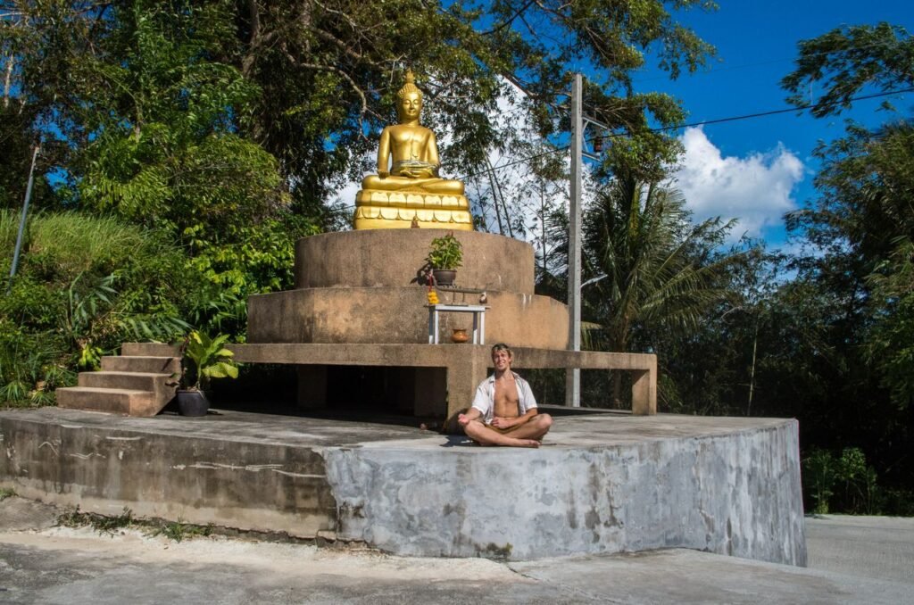 A serene Buddha statue in Phuket with a man meditating nearby. Peaceful outdoor setting.