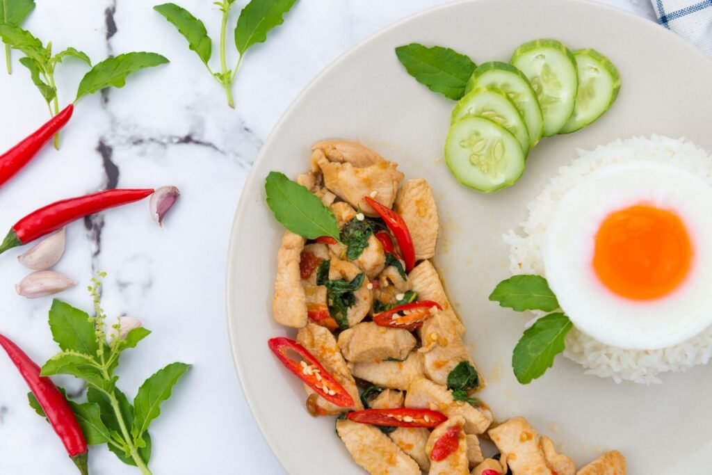 Tasty Thai basil chicken with rice, fried egg, red chili, and cucumber slices on a plate.