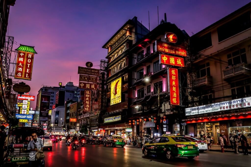 Colorful illuminated signs light up Bangkok's Chinatown street at dusk, showcasing urban life and culture.