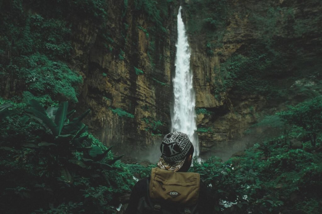 Person admiring a tall waterfall in a lush, green forest, conveying adventure and exploration.