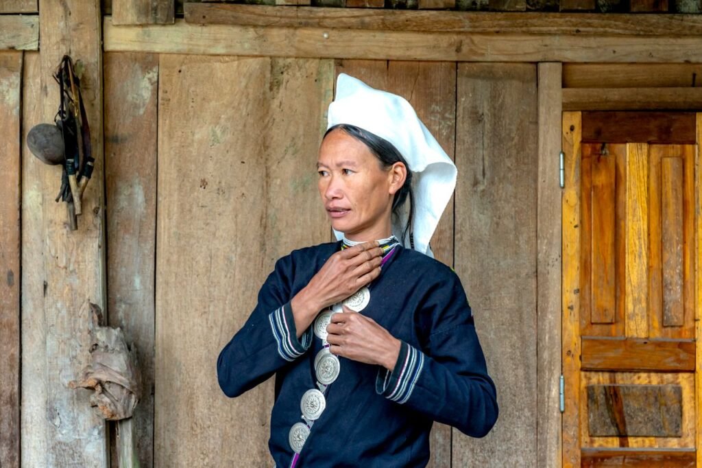 A woman stands outdoors wearing atraditional clothing and headscarf, set against a wooden wall.