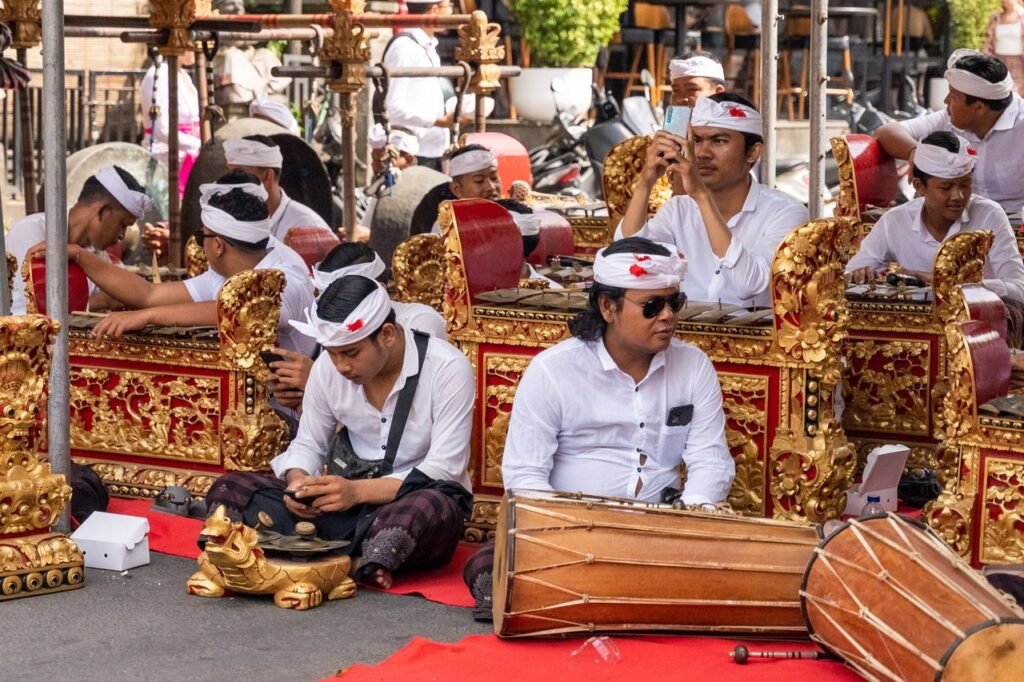 Balinese musicians perform traditional gamelan music outdoors, wearing cultural attire.