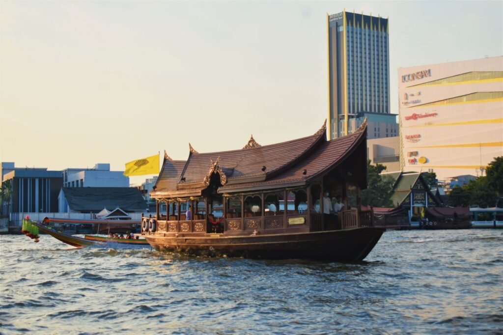 A traditional Thai boat sails on the Chao Phraya River with Bangkok's skyline in the background at dusk.