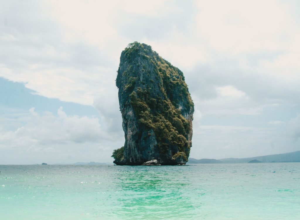 A striking limestone island emerges from the clear waters of Thailand, under a cloudy sky.
