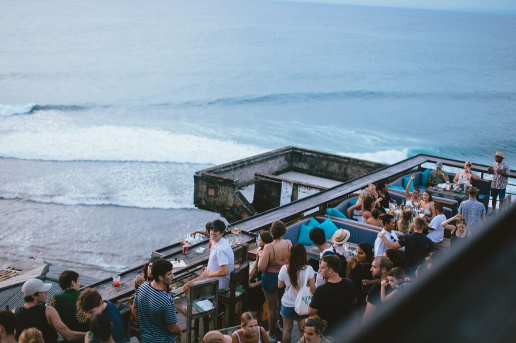 Lively outdoor bar scene by the ocean with a crowd enjoying drinks and scenic sea views.