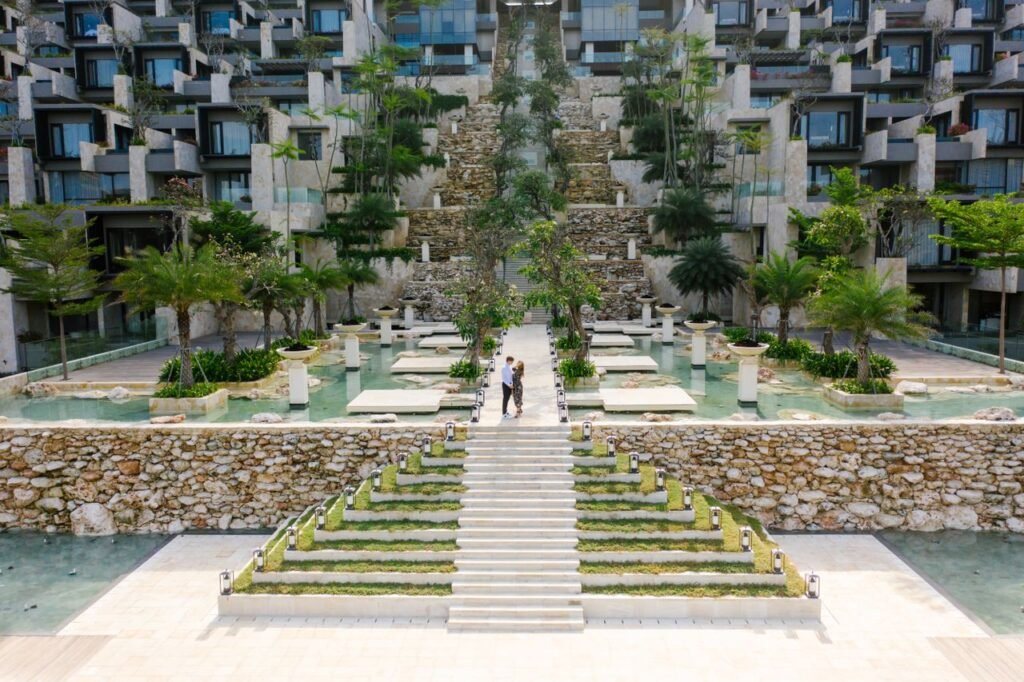 A couple walks through a modern resort with lush gardens and water features, showcasing luxurious architecture.