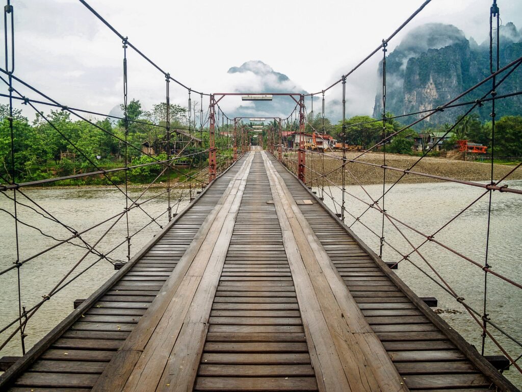 Wooden suspension bridge over river with misty mountains in Vang Vieng, Laos.