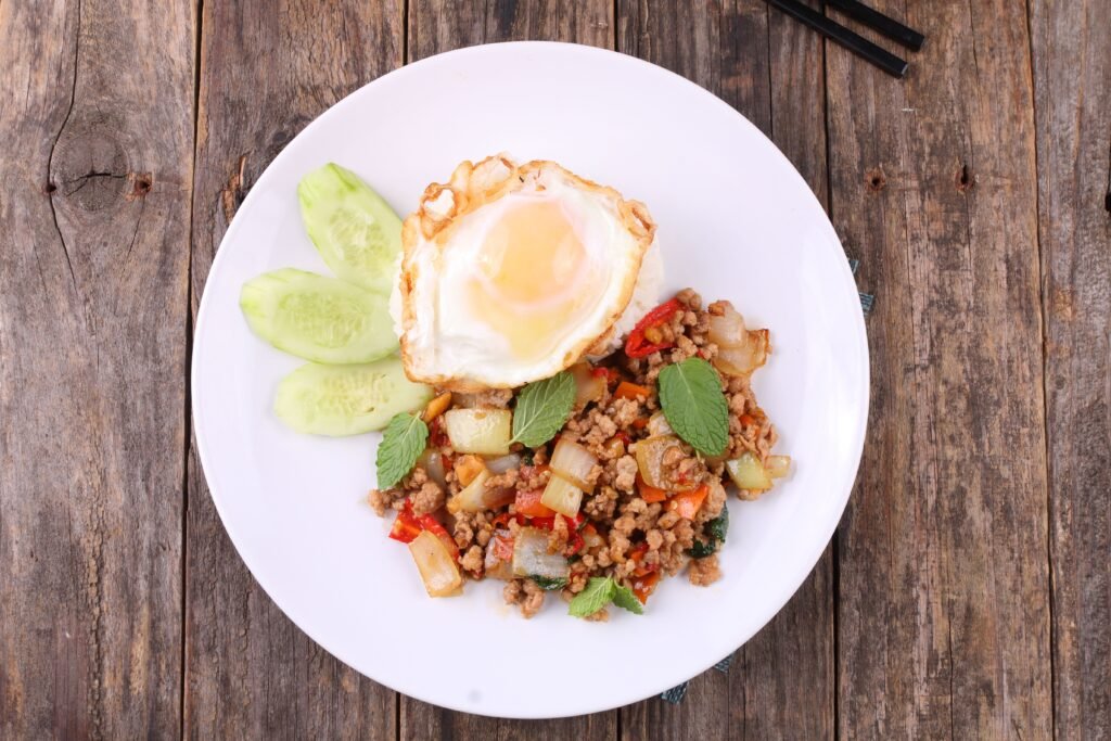 Thai basil stir-fry with minced meat, vegetables, cucumber slices, and fried egg on a wooden table.