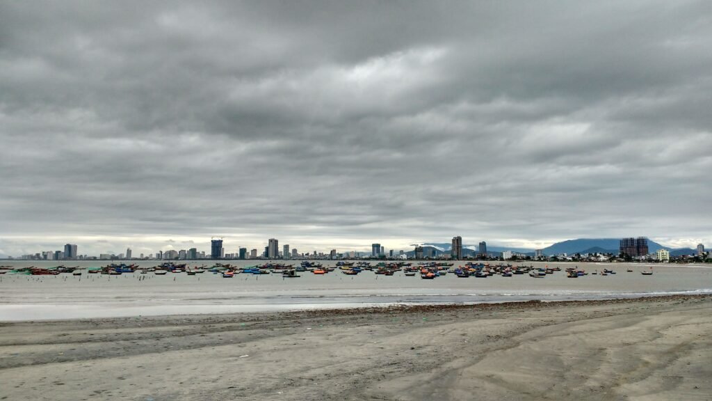 A serene view of Da Nang's skyline with fishing boats and a sandy beach under a cloudy sky.